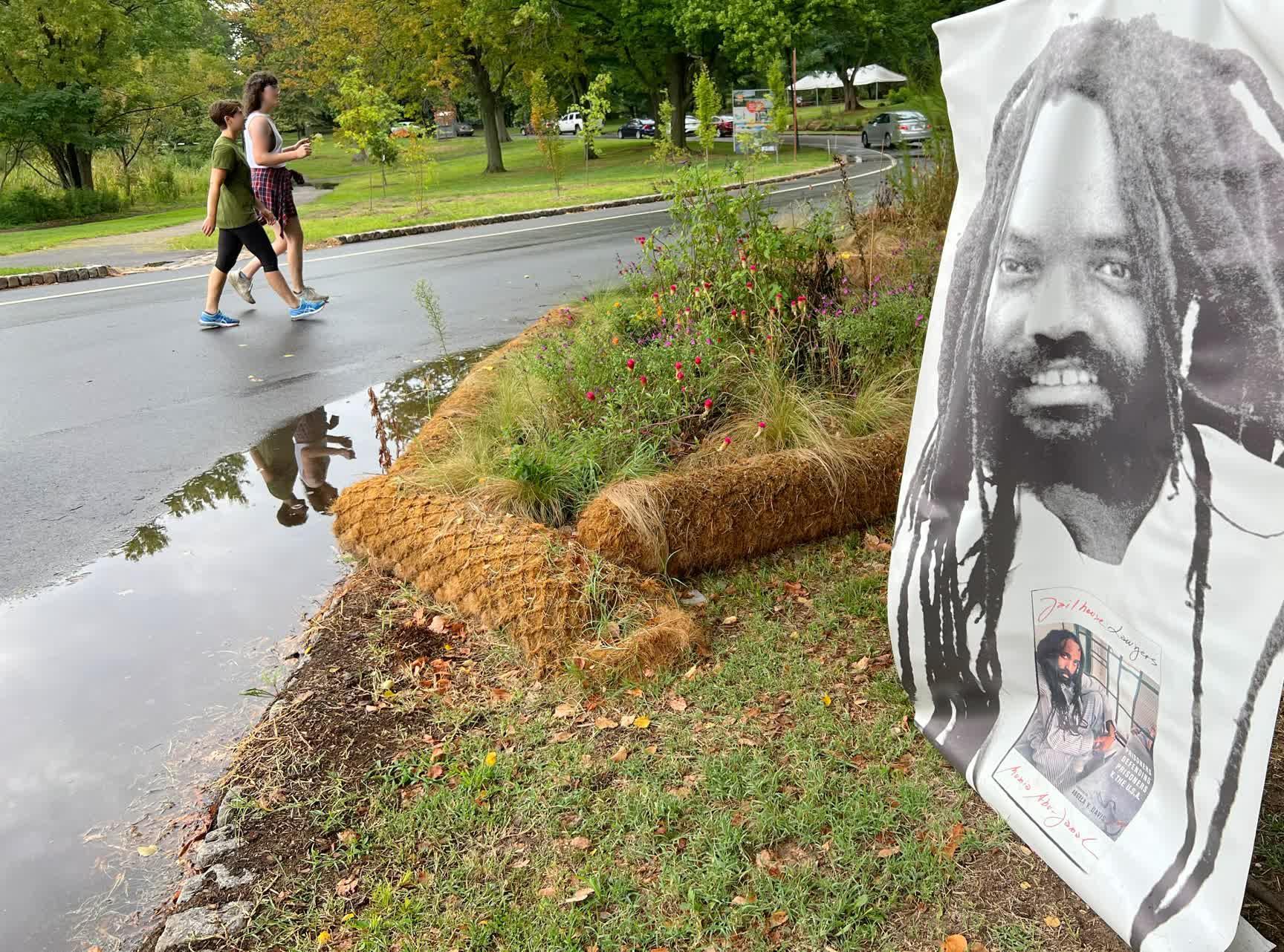 Two people walking down the road past a large banner with an image of Mumia Abu-Jamal.