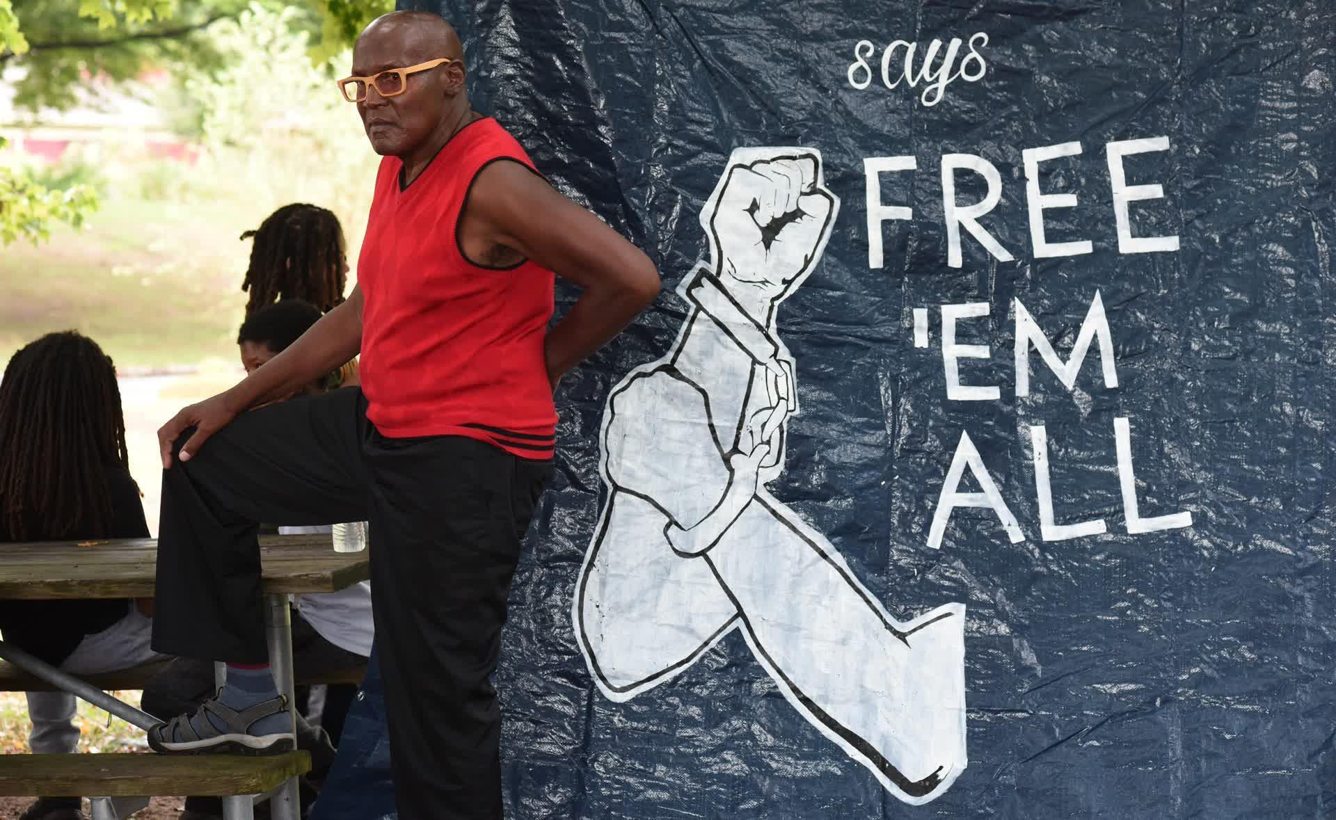 Ojore Lutalo standing next to picnic table and large banner that says free em all.