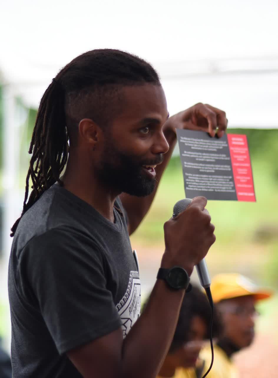 Side view of Brother O. speaking into a microphone while holding a palm card with information about MXGM.