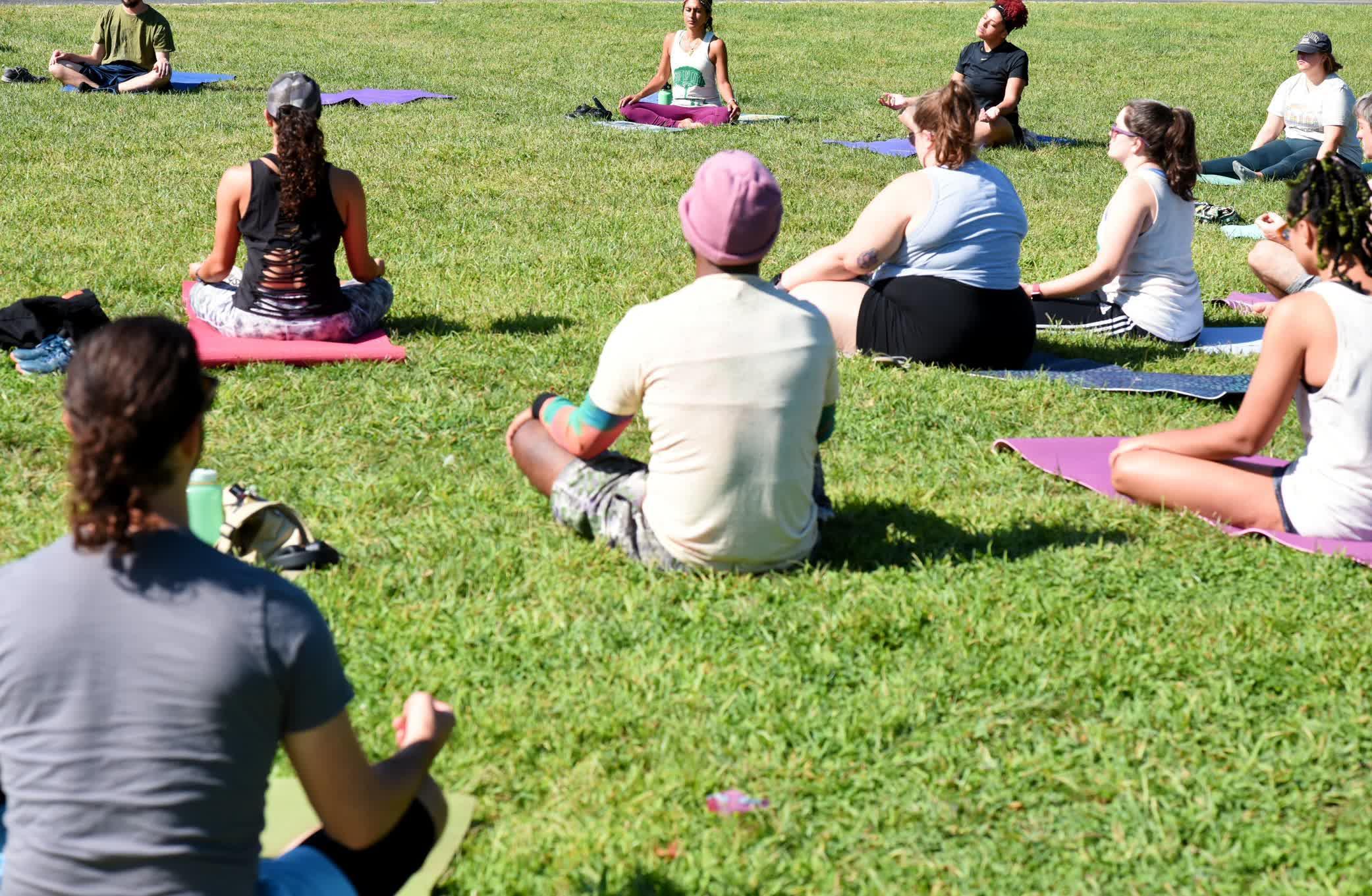 Close-up of people sitting in a circle on grass field with legs crossed and arms out doing Yoga.