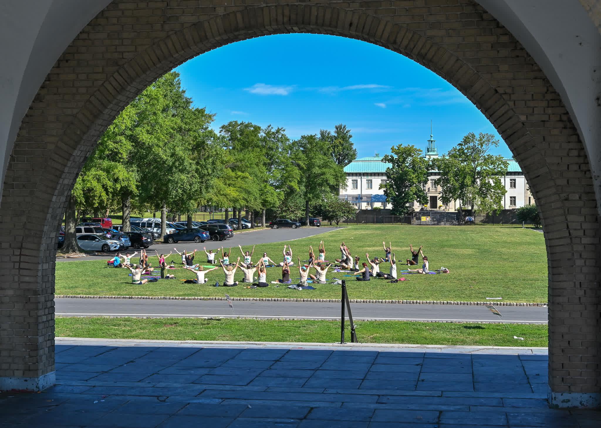 Distant view from under brick arch structure of people doing Yoga with legs crossed and arms in the air.